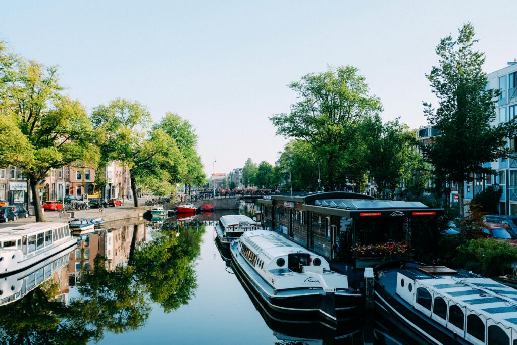 pexels-photo-3884462-3884462 Modern cruise boats moored on calm canal reflecting green trees and buildings on sunny day